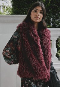 Weinrotes Kunstfellweste mit zotteliger Textur, getragen über einer dunkel gemusterten Bluse mit langem, weitem Ärmel. Schwarze gesteppte Tasche sichtbar.