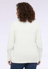 Maglione bianco lavorato a maglia con maniche lunghe, caratterizzato da dettagli in frange sui polsini. Il design è semplice e strutturato, abbinato a jeans scuri.