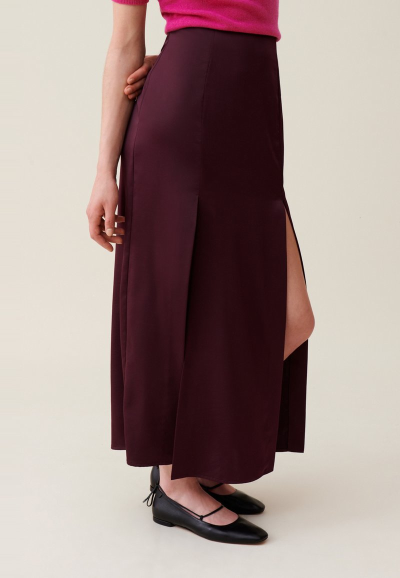 Gonna maxi bordeaux in tessuto morbido con uno spacco laterale. Abbinata a ballerine nere con un design a cinturino. Silhouette semplice ed elegante.