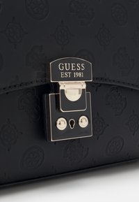 Svart läderhandväska med prägling, med lås i guldton märkt "GUESS EST 1981" och två runda nitar ovanf�ör nyckelhålet.