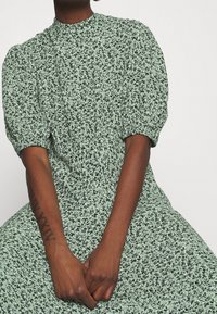 Grön mönstrad klänning med puffärmar och hög krage, gjord av ett lätt tyg med en blommönster i svart.