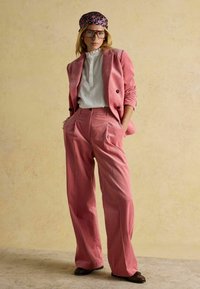 Rosa Cordhosenanzug mit weitem Hosenbein und strukturiertem Blazer, kombiniert mit einer weißen Bluse und einem bunten, gemusterten Haarband.