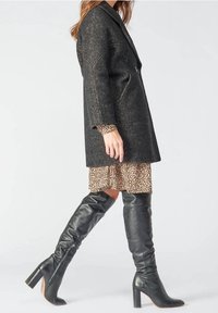 Cappotto lungo nero con texture e design sartoriale, indossato sopra un vestito leopardato bianco e nero. Abbinato a stivali neri in pelle al ginocchio.