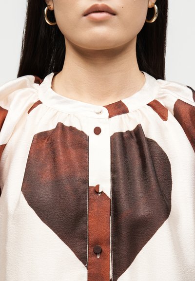 Γυναίκα που φοράει μια κρεμ μπλούζα με μεγάλο καφέ σχέδιο καρδιάς και κουμπιά καλυμμένα με ύφασμα, σε συνδυασμό με χρυσούς κρίκους σκουλαρίκια.