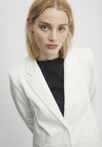 Bílý blazer s ostrými klopami a dvěma předními kapsami, nošený přes přiléhavý černý top. Modelka má světlé hnědé vlasy a přirozené pihy.
