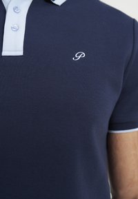 Polo bleu marine avec col bleu clair et patte de boutonnage, lettre P blanche brodée sur la poitrine, porté par une personne.