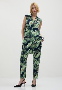 Combinaison sans manches en bleu marine avec un imprimé de feuilles tropicales vertes, comportant des poches et une coupe décontractée, associée à des escarpins noirs à bouts pointus.