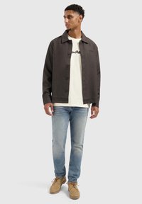 Donkergrijze button-up jas over een witte T-shirt, gedragen met lichtblauwe jeans en beige schoenen. Bevat een klassieke kraag en minimalistische accenten.