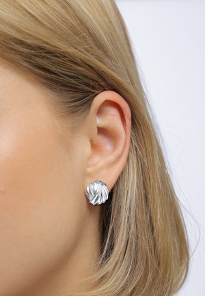 Boucles d'oreilles en métal argenté avec un design texturé et ondulé évoquant des pétales, montées sur une petite base ronde, présentées à côté d'une oreille.