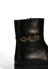Zwarte leren enkellaars met een glanzende afwerking, voorzien van een goudkleurige gespdetail en een elegante silhouet. Zij rits voor gemakkelijke aan- en uittrekken.