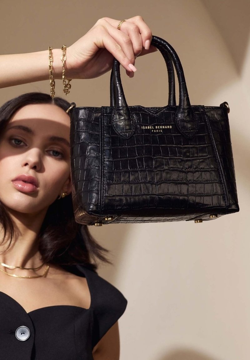 Zwarte handtas met krokodillenstructuur, korte handgrepen en gouden hardware. Heeft een rechthoekige vorm en een gladde, glanzende textuur.
