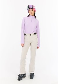 Svetlooranžový dlhý zipsový top, béžové nohavice s vreckami a čierne lyžiarske topánky. Model má na sebe ružovú a fialovú čiapku a okuliare.