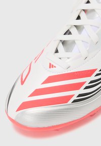 Scarpone da calcio bianco con strisce rosse, dettagli argentati e superficie testurizzata. Presenta lacci e branding "MESSI" sul lato.