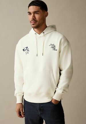 Cream hoodie with front pocket, drawstring hood, and navy floral embroidery on the left chest. Features ribbed cuffs and hem.