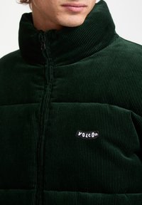 Dark green corduroy puffer jacket with a high collar, featuring vertical texture and a black fabric logo patch on the left chest.