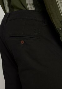 Pantaloni neri con una texture liscia, dotati di una tasca posteriore con un bottone marrone e un discreto logo ricamato. La vita è semplice.