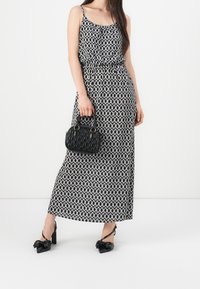 Maxikleid in Schwarz-Weiß mit Muster, dünnen Trägern und elastischer Taille, kombiniert mit einer kleinen schwarzen, strukturierten Handtasche und Schuhen mit klobigem Absatz.