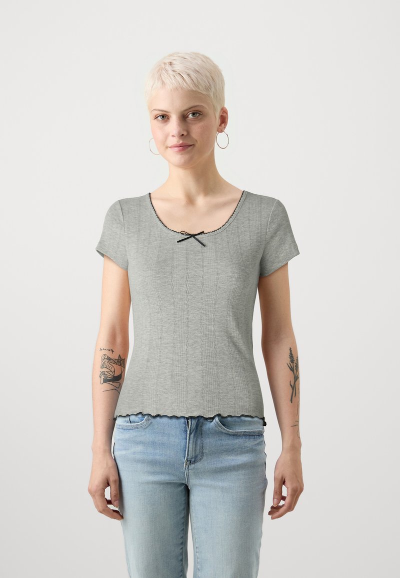 Top grigio a maniche corte con orlo a scollatura, dotato di un accento a fiocco nero sul collo e tessuto textured. Indossato con jeans blu.