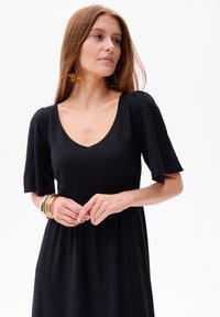 Schwarzes, knielanges Kleid mit V-Ausschnitt, kurzen Puffärmeln und lockerer Passform. Glatte Textur; kombiniert mit goldenen Armreifen und Blattohrringen.