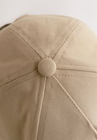 Cappellino beige con corona rotonda e dettaglio con bottone sulla parte superiore, realizzato in tessuto strutturato con cuciture visibili lungo le giunture.