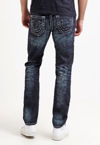 Mörkblå denimjeans med en urblekt finish, kontrasterande sömmar och distinkta bakfickor med en unik design. Bärs med vita skor.