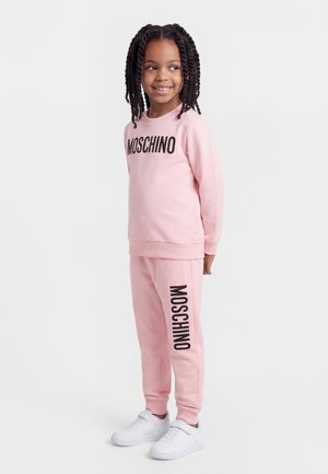 Jeune fille aux cheveux tressés portant un sweat-shirt Moschino rose assorti et un pantalon de survêtement, avec des baskets blanches, souriant et regardant sur le côté.