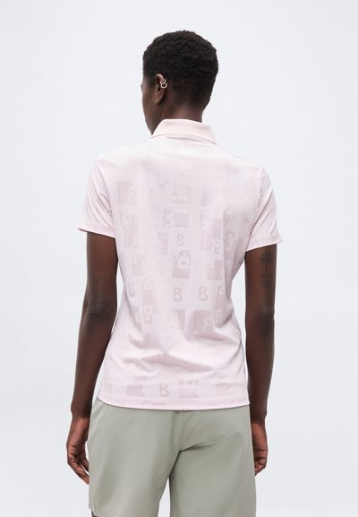 Personne aux cheveux courts et bouclés portant un polo rose clair avec des carrés à motifs subtils et un short gris clair, vue de dos.