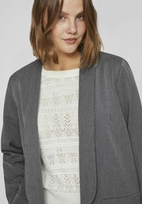 Blazer gris au toucher lisse, avec un design épuré et une ouverture à l'avant. En dessous, un pull en tricot blanc avec un motif floral.