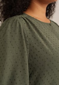 Blusa verde con una morbida texture, caratterizzata da un motivo a pois e maniche leggermente arricciate. Il décolleté include un piccolo dettaglio con un bottone.