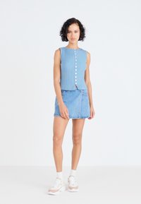 Lichtblauw geribbeld gebreid mouwloos topje met witte knopen, gecombineerd met een denim minirok. Model draagt witte sneakers met beige accenten.