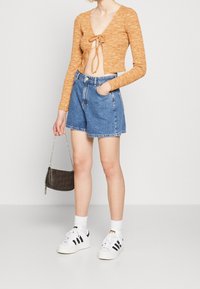 Orange texturerad crop top med knytframsdetail, i kombination med höga blå denimshorts. Vita sneakers med svarta ränder och en liten handväska.