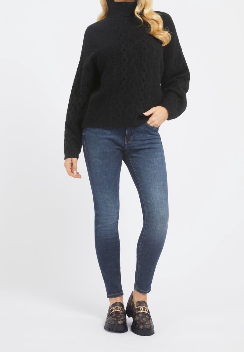 Schwarzer Kabelstrickpullover mit hohem Kragen, kombiniert mit blauen Skinny-Jeans. Die Schuhe besitzen goldene Hardware und ein dunkles, gemustertes Design.