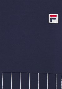 Vêtement bleu marine avec un petit logo carré blanc présentant un motif rouge et noir, et des rayures blanches verticales en bas.