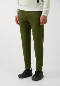 Olivgrüne Sweatpants mit elastischem Bund und Bündchen an den Knöcheln; der Stoff ist glatt; kombiniert mit schwarzen Sportschuhen.