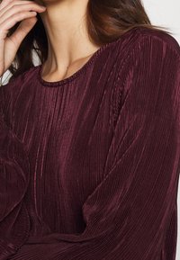 Burgunderfarbene Langarmbluse mit gerippter Textur, runder Halslinie und gerafften Details an den Ärmeln. Weiches, leichtes Material.
