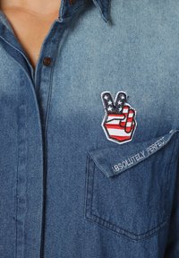 Camisa de mezclilla en color azul claro con collar puntiagudo. Presenta un parche de símbolo de la paz en rojo, blanco y azul y bordado "ABSOLUTAMENTE PERFECTO".