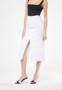 Gonna midi in denim bianco con uno spacco frontale, vita alta e una silhouette aderente. Abbinata a mule con tacco aperto nero e bianco.
