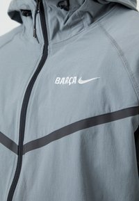 Ανοιχτό γκρι μπουφάν με φερμουάρ, μαύρες λεπτομέρειες, λογότυπα "BARÇA" και Nike σε λευκό χρώμα. Κατασκευασμένο από απαλό, ελαφρύ ύφασμα.