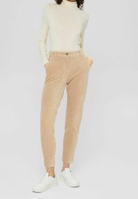 Beige kordfloddsbyxor med smal passform, med sidofickor och en enkel knappstängning, kombinerat med vita sneakers och en ljus polotröja.