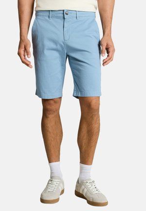 Mężczyzna ubrany w jasnoniebieskie spodenki do kolan, białe skarpetki, beżowe trampki i białą koszulę wpuszczoną w spodnie, stojący na jednolitym tle.