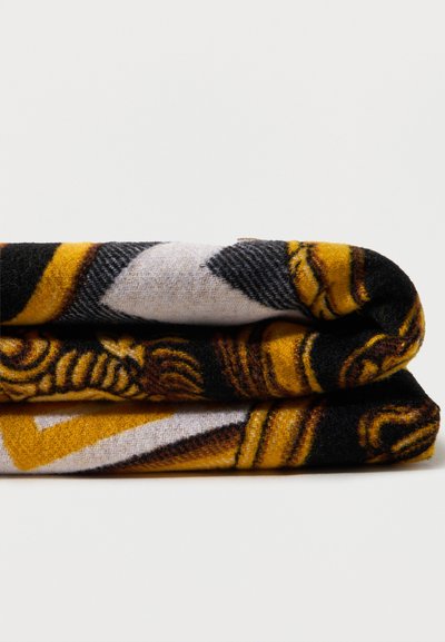 Διπλωμένη μάλλινη κουβέρτα με μαύρα, κίτρινα και λευκά γεωμετρικά και περίτεχνα σχέδια σε λευκό φόντο.