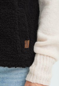 Zwarte fleece stof met een zachte textuur, gecombineerd met een geribbelde crèmekleurige mouw van een sweater. Inclusief een bruine label met "Oxmo" branding.