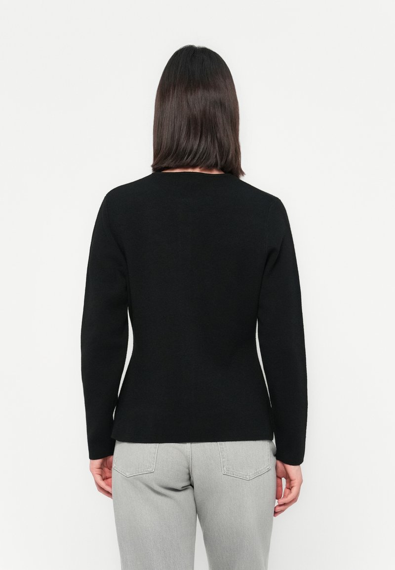 Kobieta z prostymi ciemnymi włosami, nosząca czarną bluzkę z długim rękawem i jasnoszare dżinsy, stojąca na tle białej ściany.