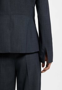 Blazer grigio scuro su misura con cuciture visibili, spacchi laterali e spalle strutturate, abbinato a pantaloni su misura coordinati.