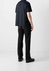 Marineblauw shirt met korte mouwen en een iets langere achterkant, gecombineerd met zwarte rechte jeans en donkere schoenen. Eenvoudig, modern ontwerp.