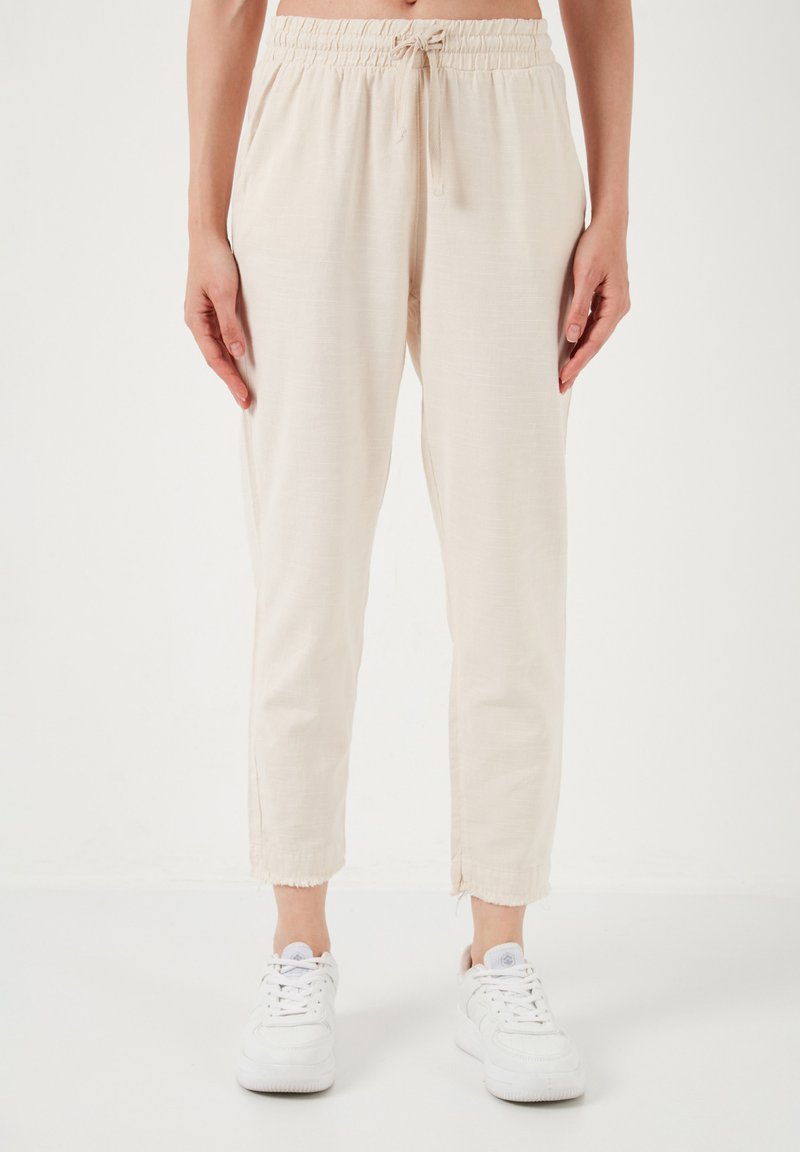 Light beige, lightweight pants with an elastic waistband and drawstring. Tapered fit with frayed hem. Paired with white sneakers.