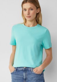 T-shirt turquoise à manches courtes avec un col rond, en tissu doux; porté avec un jean, mains dans les poches. Design simple et épuré.