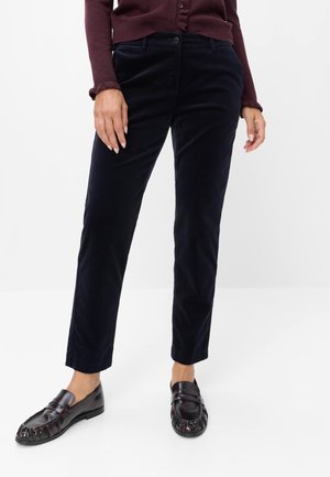 Kvinde iført sorte bukser med lige ben, mørk burgunderrød knappet sweater og sorte læderloafers, står mod en ensfarvet hvid baggrund.