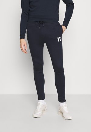 Mand iført sorte slim-fit joggers med "11°" logo på låret, sort sweatshirt, hvide sokker og beige sneakers, stående mod en ensfarvet baggrund.
