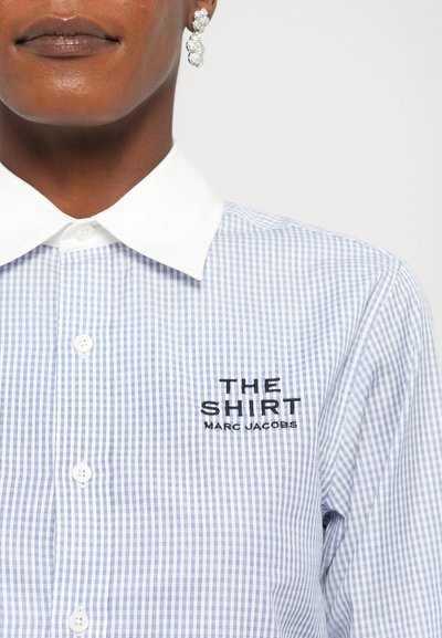 Modrá a biela kockovaná košela s bielym golierom. Na hrudi je čierny vyšívaný text "THE SHIRT MARC JACOBS". Zapínanie na gombíky.